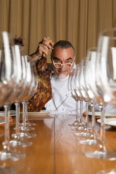 השף שאול בן אדרת אוחז בנתח בשר ומלפניו שורת כוסות זכוכית במסעדת "התרנגול הכחול"